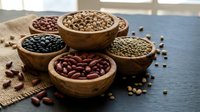 Assorted legumes and beans in wooden bowls on dark surface with burlap cloth, showcasing variety of protein-rich foods for healthy cooking and nutrition.