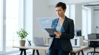 Middle-aged woman with short dark hair, Caucasian ethnicity, wearing professional business attire with blazer, holding tablet device while analyzing digital data visualization in modern office environment.