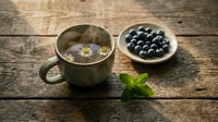 Herbal tea with chamomile flowers in rustic cup beside fresh blueberries and mint leaves on wooden surface for wellness and natural beverage concepts.