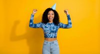 Excited young African American woman celebrating birthday with party hat raising arms in victory pose against bright yellow background.