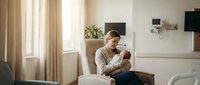Young Caucasian mother in beige sweater tenderly holding and bonding with newborn baby in bright modern living room with natural sunlight streaming through windows.