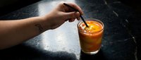 Person stirring iced cola drink with spoon in glass on dark table surface, beverage consumption and refreshment concept.