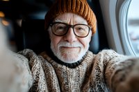 Cheerful senior Caucasian man with gray beard wearing knit beanie and sweater takes selfie while traveling by plane, showing genuine happiness and active lifestyle.