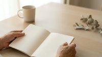 Mature adult with brown skin tone holding open blank notebook on wooden desk with coffee mug and dried eucalyptus, suitable for journaling, writing, planning, and lifestyle content.