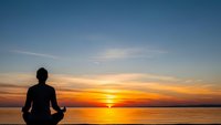 Silhouette of person meditating in lotus pose at sunset over calm water. Peaceful mindfulness and wellness concept for relaxation and spiritual practice.