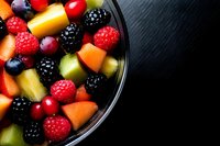 Fresh mixed fruit salad with raspberries, blackberries, blueberries, grapes and tropical fruit chunks in glass bowl on dark background. Healthy eating concept.