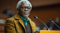 Senior African American man with glasses and mustache speaking at podium, wearing yellow tweed jacket and green shirt, expressing warmth and confidence during presentation.