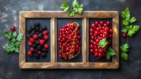 Fresh summer berries assortment in rustic wooden box compartments with blackberries, red currants, and raspberries on dark background with parsley leaves.