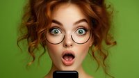 Young woman with curly red hair and round glasses expressing shock while looking at smartphone screen against green background. Emotional portrait capturing surprise moment.