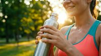 Woman athlete in coral sports top holding water bottle during outdoor workout in sunny park, staying hydrated during exercise routine.