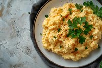 Creamy scrambled eggs garnished with fresh parsley on white plate against rustic concrete background. Classic breakfast dish with pepper seasoning.