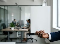 Modern professional holding blank white card in contemporary office workspace with colleagues collaborating in background, suitable for corporate messaging and business communication campaigns.