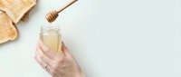 Hand holding glass jar of golden honey with wooden dipper above, toasted bread slices nearby on white background, suitable for food, wellness and cooking content.