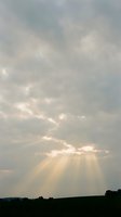 Dramatic sky with golden sunrays breaking through layered clouds above a dark landscape horizon. Crepuscular rays create ethereal light beams for spiritual, inspirational, or weather-related projects.