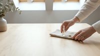 Woman cleaning wooden table surface with white cloth in bright modern kitchen interior with natural lighting and minimalist design elements.