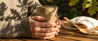 Hands in cozy knit sweater holding warm ceramic mug on wooden table with autumn leaves and soft sunlight shadows creating peaceful morning atmosphere.