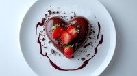 Heart shaped chocolate mousse dessert garnished with fresh strawberries and chocolate shavings on white plate with berry sauce drizzle, elegant restaurant presentation.