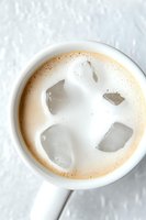 Creamy iced coffee drink in white ceramic mug with ice cubes floating on top, photographed from above on light textured background. Fresh morning beverage.