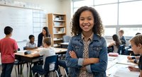 Young mixed-race female teacher with curly hair smiling confidently in modern classroom with diverse students working at desks in background during school day.