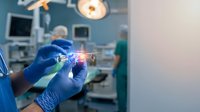 Surgeon in blue gloves holding surgical instrument with glowing light effect in operating room with medical equipment and staff in background.