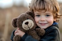 Smiling Caucasian toddler boy with blue eyes hugging brown teddy bear, wearing dark sweater. Close up portrait showing genuine happiness and childhood innocence.