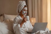 Woman in bathrobe with facial mask enjoying coffee while working on laptop in bed during morning self-care routine.