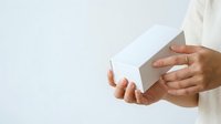 Person of medium skin tone holding white rectangular box with both hands against neutral background, suitable for product packaging, unboxing, delivery, and retail concepts.