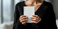Woman in black sweater holding white book with blank cover, showcasing elegant jewelry and manicured nails in soft natural light.