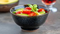 Fresh avocado and cherry tomato salad garnished with basil leaves in black ceramic bowl, served on dark stone surface with wine glass in background.