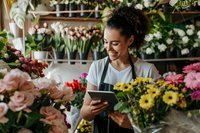 Young mixed race female florist with curly hair in apron using digital tablet while working among colorful fresh flowers in modern flower shop. Small business owner.