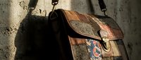 Vintage leather messenger bag with woven textile details hanging against textured concrete wall. Classic brown satchel with ethnic pattern accents for professional use.