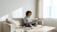 Young African American woman working remotely on laptop computer while sitting comfortably on modern beige sofa in bright living room with natural sunlight.