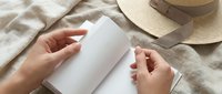 Woman hands holding blank white card mockup on beige linen fabric with straw hat. Clean minimal scene for branding, invitation or greeting card design.