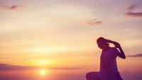 Silhouette of contemplative woman sitting during golden hour sunset with warm pink and orange sky colors creating peaceful meditation moment.