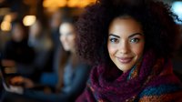 Young African American woman with natural curly hair smiling warmly at camera, wearing colorful knit scarf. Soft bokeh background creates cozy evening atmosphere.