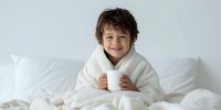 Young boy with tousled brown hair wrapped in white blanket holding warm mug in cozy bedroom setting. Child enjoying hot beverage on bed with soft linens.