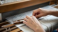 Skilled artisan hands weaving natural linen fabric on traditional wooden loom, showcasing handcraft textile production and sustainable fiber arts manufacturing process.