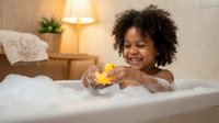 Happy African American toddler girl playing with yellow rubber duck toy in bubble bath at home. Child enjoying bath time in cozy bathroom setting.
