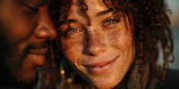 Close-up portrait of young woman with freckles and curly hair smiling at camera while man leans close in warm golden light.