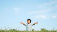 Young boy with curly dark hair wearing checkered shirt with arms spread wide in joyful celebration outdoors in meadow with wildflowers and blue sky.