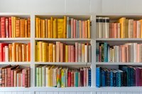 Colorful books arranged by hue on white wooden shelves create rainbow spectrum from red to blue, organized library collection for home or bookstore interior.