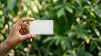 Hand holding blank white card against blurred green foliage background, suitable for business card mockups, branding presentations, and marketing material designs.