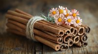 Bundle of aromatic cinnamon sticks tied with twine and decorated with small pink daisy flowers on rustic wooden background, cozy kitchen spice setup.