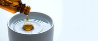 Golden oil pouring from amber bottle into white ceramic bowl creating ripples on clean background for cooking and wellness applications.