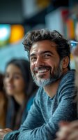 Happy mature Hispanic man with curly gray hair and beard smiling warmly in casual sweater at social gathering with friends in background.