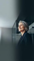 Mature woman with gray hair wearing dark blazer standing in modern office building with glass windows, looking away thoughtfully in professional corporate environment.