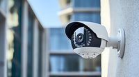 Modern security surveillance camera mounted on white building wall with infrared LED lights for night vision monitoring, focused view against blurred urban background.