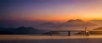 Wooden table and chair on terrace overlooking misty mountains and lake at golden sunrise, serene landscape for travel and wellness design projects.