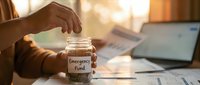 Person saving money in emergency fund jar at home office desk with financial documents and laptop for budget planning and financial security.