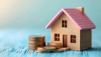 Small model house with pink roof next to stack of gold coins on blue wooden surface, representing real estate investment and property finance concept.